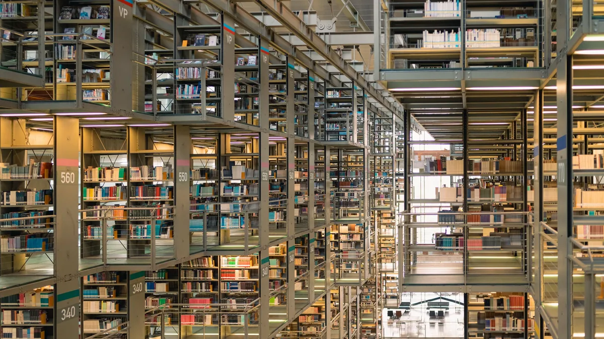 A large library with multiple levels of shelving units, each containing numerous books, and a staircase leading to the upper levels.