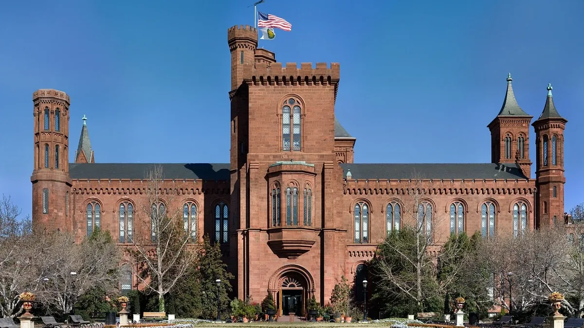 The Smithsonian Castle, a red brick building with a flag on top, situated in a park-like setting with trees and grass.