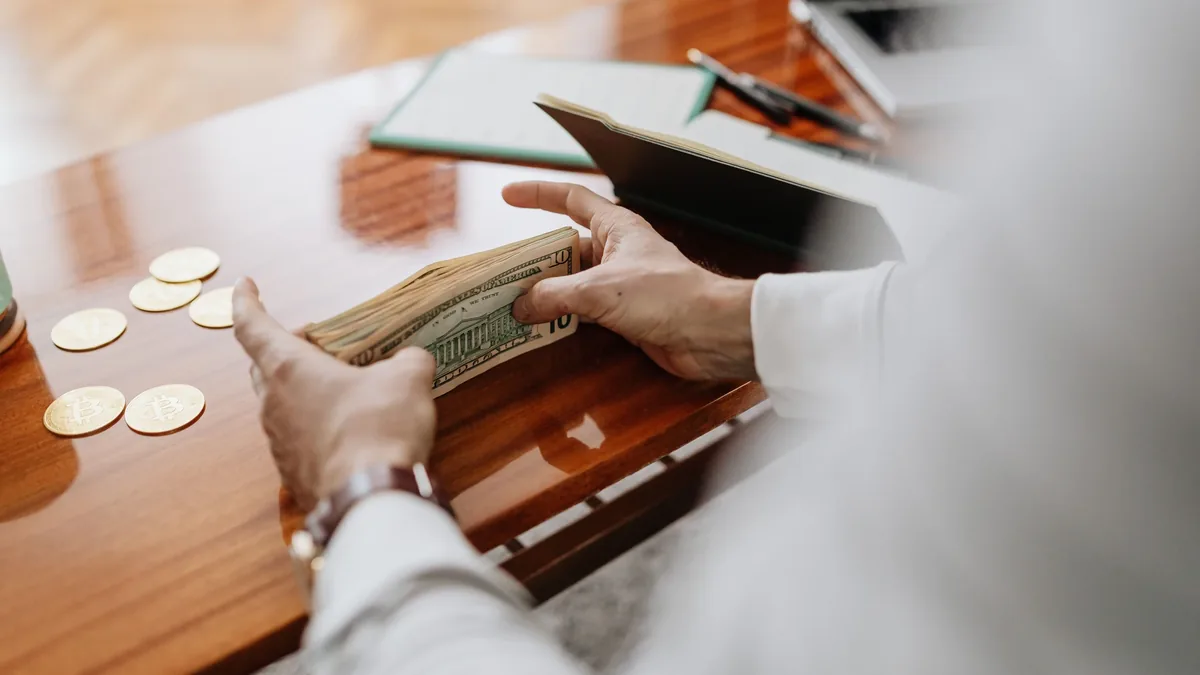 A person is visible from the chest down, seated at a table with a glossy surface, holding a stack of cash in their hands.